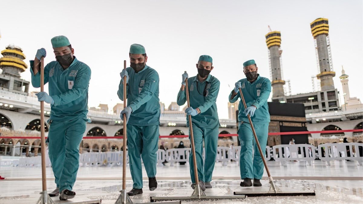 Workers cleaning the white marble courtyard of Masjid al-Haram with the clock tower and hotel towers behind