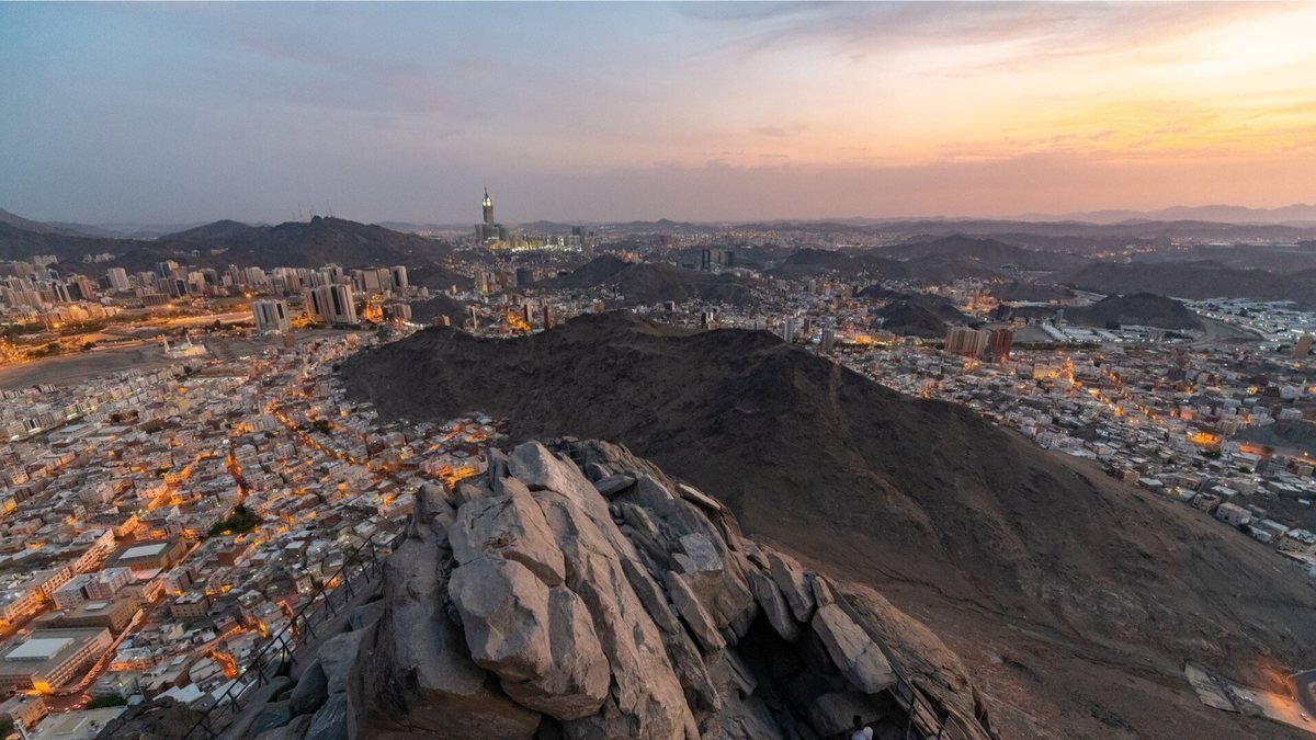 Makkah at sunset seen from a rocky hillside, with the clock tower rising above the city in the distance