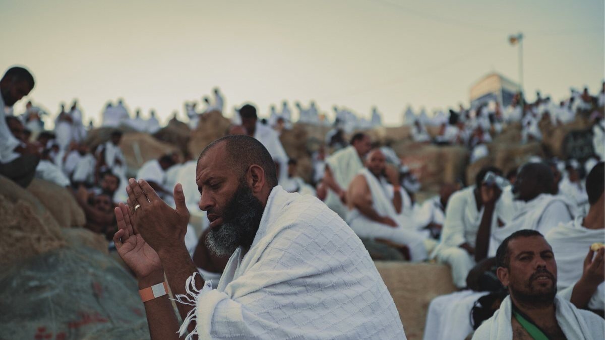 A pilgrim in white Ihram sheets raising his hands in supplication among a seated crowd