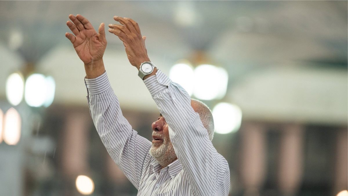 An older pilgrim in white raising both hands in supplication inside a mosque