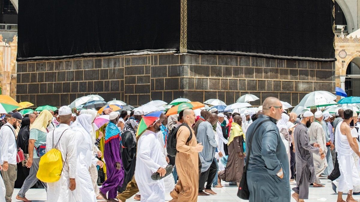 Pilgrims walking in tawaf around the Kaaba in daylight, several carrying umbrellas against the sun